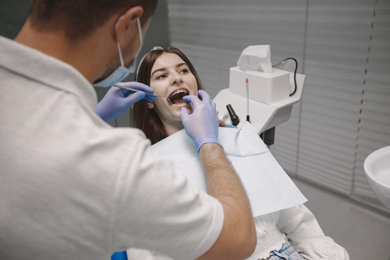 Female patient with braces has dental examination at dentist office. Woman wearing white clothes. Male dentist treats girl.